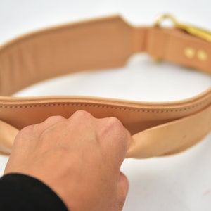 Op de afbeelding: Een bruine leren riem met een gouden gesp. De riem is dubbelgevouwen en een hand houdt de twee uiteinden bij elkaar.