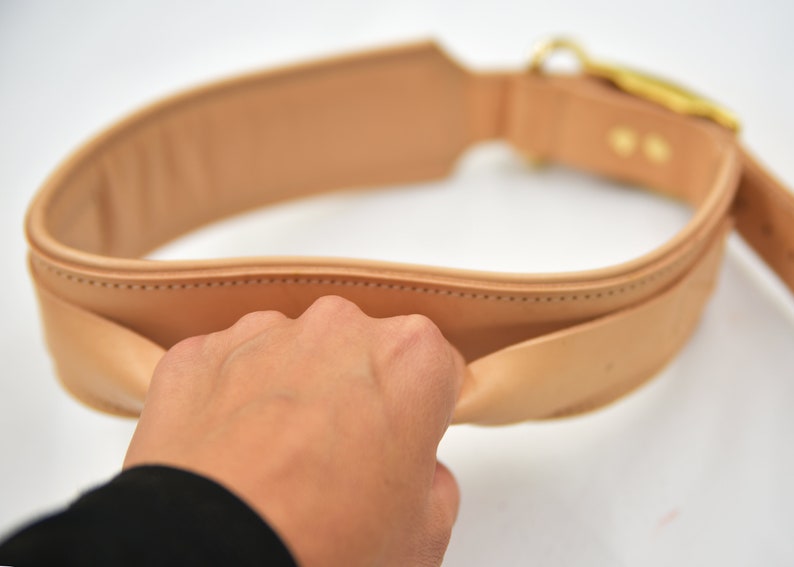 Op de afbeelding: Een bruine leren riem met een gouden gesp. De riem is dubbelgevouwen en een hand houdt de twee uiteinden bij elkaar.