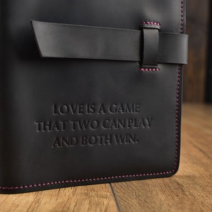 以下が含まれることがあります： ピンクのステッチと革のストラップ留め具が付いた黒い革のジャーナル。ジャーナルには、「愛は二人でできるゲームで、どちらも勝てる」という引用が前面に刻印されています。