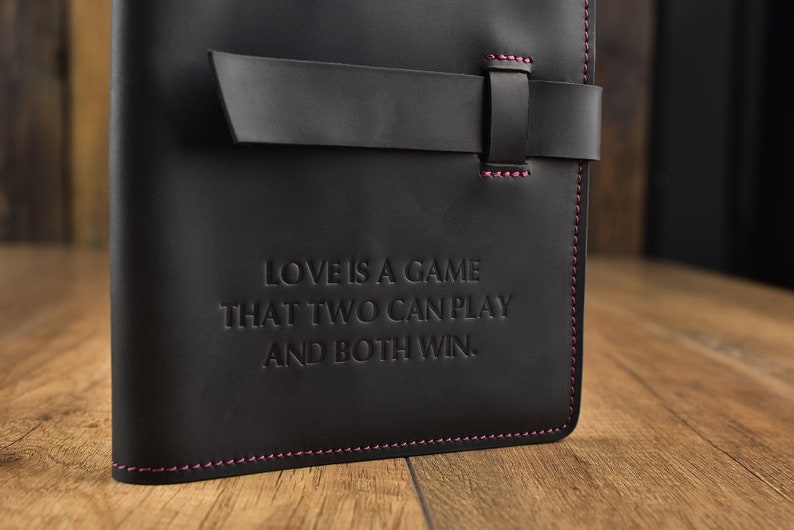 以下が含まれることがあります： ピンクのステッチと革のストラップ留め具が付いた黒い革のジャーナル。ジャーナルには、「愛は二人でできるゲームで、どちらも勝てる」という引用が前面に刻印されています。