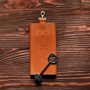 May include: A brown leather key holder with a silver key ring and two small hearts embossed on the front. A silver key is attached to the key ring.
