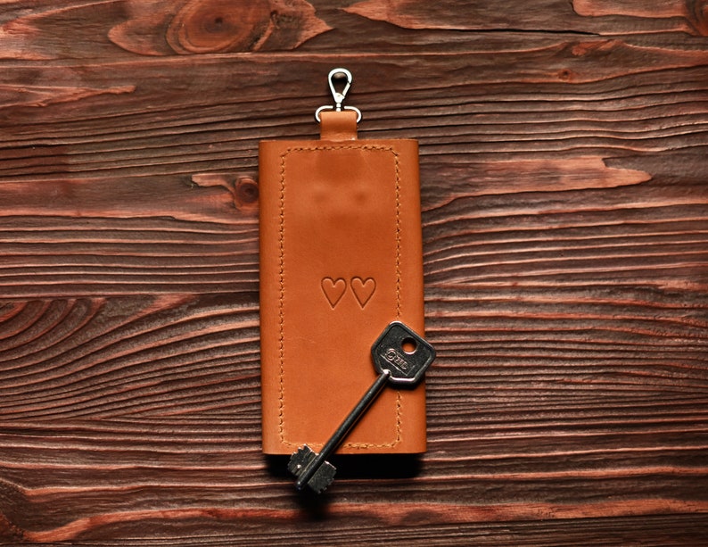 May include: A brown leather key holder with a silver key ring and two small hearts embossed on the front. A silver key is attached to the key ring.