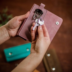 May include: A pink leather key holder with a silver key chain and keys. The key holder is being held by a person's hands. There are three other key holders in the background, one is turquoise, one is brown, and one is orange.