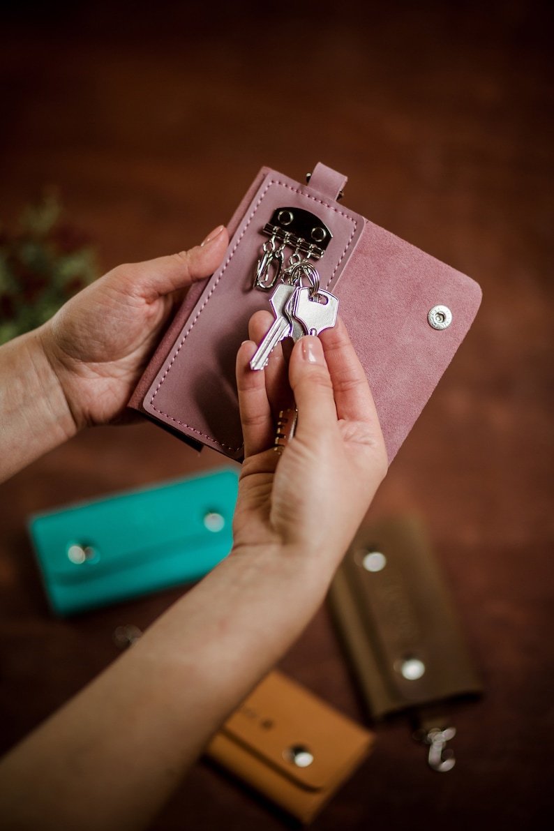 May include: A pink leather key holder with a silver key chain and keys. The key holder is being held by a person's hands. There are three other key holders in the background, one is turquoise, one is brown, and one is orange.