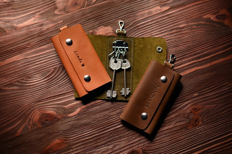 May include: Two brown leather keychains with the names "Linda" and "Brandon" embossed on them. The keychains are on a wooden surface and one keychain has a set of keys attached.