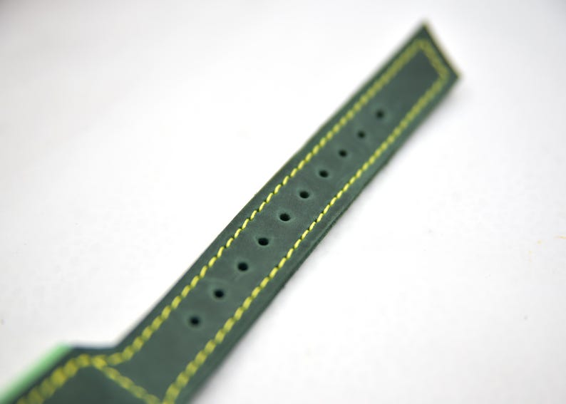 Puede incluir: Una correa de reloj de color verde oscuro con acabado mate y costuras de contraste en color amarillo brillante a lo largo de los bordes. La correa incluye una fila &uacute;nica de orificios de ajuste circulares espaciados uniformemente.