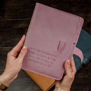 May include: A mauve leather journal with the embossed text "NATURE IS PAINTING FOR US, DAY AFTER DAY, PICTURES OF INFINITE BEAUTY." The journal has a strap closure and is held over other leather items.