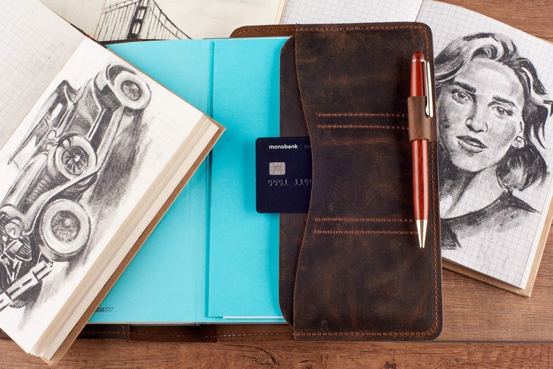 May include: A brown leather notebook cover with a pen holder and a card slot. The cover has a stitched design and is open to reveal a blue notebook page. A credit card is inserted in the card slot. The notebook cover is on a wooden surface.