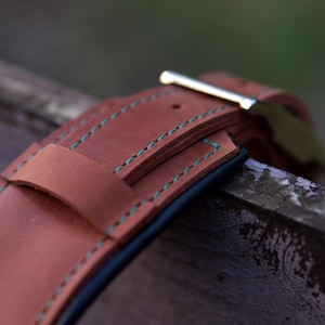 Puede incluir: Una correa de reloj de cuero color canela con costuras en contraste de color verde azulado a lo largo de los bordes. La correa incluye una trabilla de cuero y una hebilla de metal plateado pulido, con una construcci&oacute;n en capas visible.