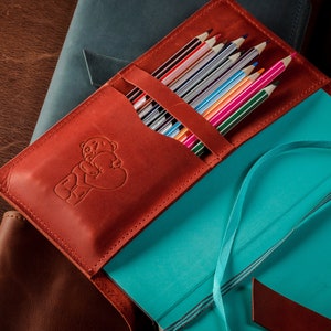 May include: A brown leather journal with a blue cover and a pocket for colored pencils. The journal has a bear design on the front and a blue ribbon bookmark.