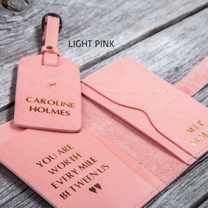 Puede incluir: Conjunto de portapasaporte y etiqueta de equipaje de cuero rosa claro con letras doradas. El portapasaporte tiene el texto "You are worth every mile between us" y la etiqueta de equipaje tiene el texto "Caroline Holmes".