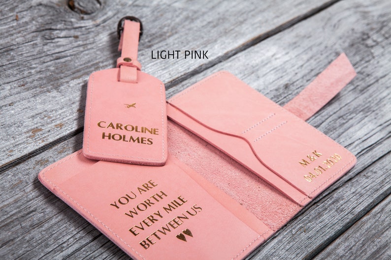 Puede incluir: Conjunto de portapasaporte y etiqueta de equipaje de cuero rosa claro con letras doradas. El portapasaporte tiene el texto "You are worth every mile between us" y la etiqueta de equipaje tiene el texto "Caroline Holmes".