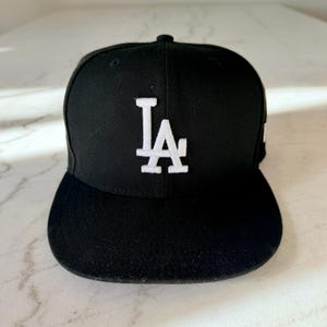 May include: Black baseball cap with a flat brim. The cap features the white embroidered letters "LA" on the front. The hat is a classic style, suitable for casual wear and sports.