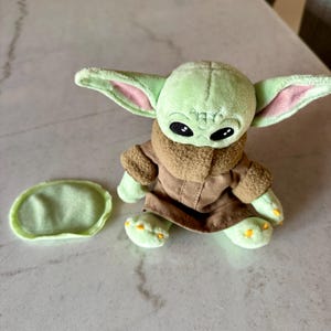Pu&ograve; includere: Un peluche verde raffigurante un personaggio con grandi orecchie e occhi neri. Indossa un cappotto marrone con un colletto peloso. Un piccolo pezzo verde di forma ovale &egrave; accanto al giocattolo. Il giocattolo &egrave; su una superficie chiara.