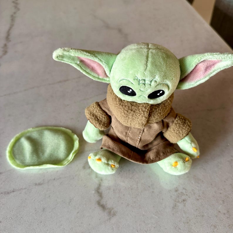 Pu&ograve; includere: Un peluche verde raffigurante un personaggio con grandi orecchie e occhi neri. Indossa un cappotto marrone con un colletto peloso. Un piccolo pezzo verde di forma ovale &egrave; accanto al giocattolo. Il giocattolo &egrave; su una superficie chiara.