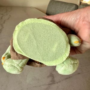 Pu&ograve; includere: Un giocattolo a forma di tartaruga in peluche, nei toni del verde e del marrone. La tartaruga ha un corpo verde chiaro, arti verde scuro e un guscio marrone. I piedi hanno dettagli gialli.