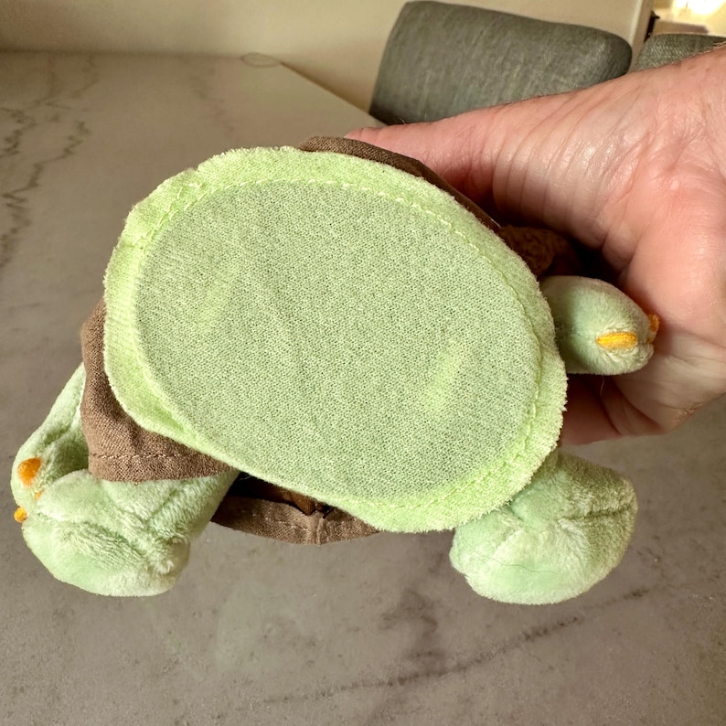 Pu&ograve; includere: Un giocattolo a forma di tartaruga in peluche, nei toni del verde e del marrone. La tartaruga ha un corpo verde chiaro, arti verde scuro e un guscio marrone. I piedi hanno dettagli gialli.