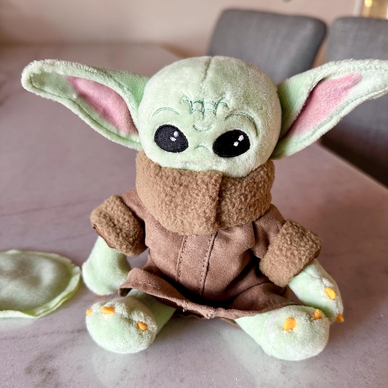 Pu&ograve; includere: Una bambola di peluche di un personaggio con grandi occhi neri, pelle verde e grandi orecchie. La bambola indossa un cappotto marrone con un colletto marrone. La bambola &egrave; seduta su una superficie di colore chiaro.