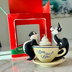 May include: A ceramic ornament featuring Pepe Le Pew and Penelope Pussycat. The characters are positioned around a coffee cup with the words "Love You A Latte!" The ornament has a white whipped cream topping and a gold loop for hanging. The ornament is in a red box.
