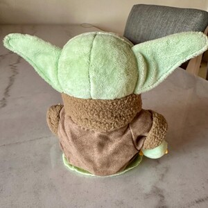 Pu&ograve; includere: Una bambola di peluche di un personaggio con grandi orecchie a punta e un incarnato verde chiaro. La bambola indossa una veste marrone con un colletto sfocato. La schiena della bambola &egrave; rivolta verso la fotocamera.