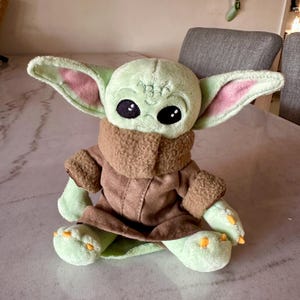 Pu&ograve; includere: Una bambola di peluche di un personaggio con grandi orecchie, pelle verde e grandi occhi neri. La bambola indossa un cappotto marrone e una sciarpa marrone chiaro. La bambola &egrave; seduta su una superficie di marmo.