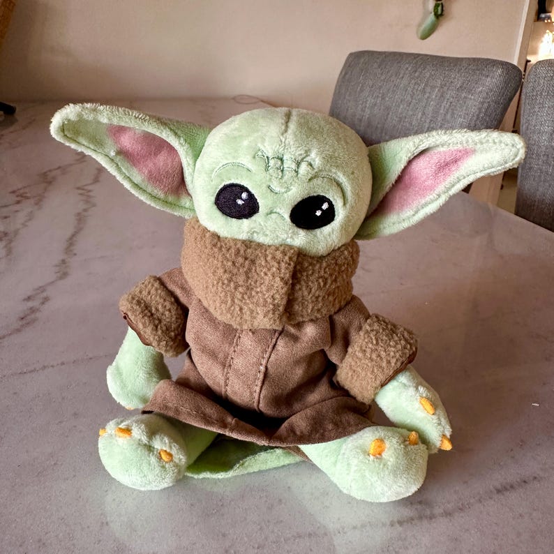 Pu&ograve; includere: Una bambola di peluche di un personaggio con grandi orecchie, pelle verde e grandi occhi neri. La bambola indossa un cappotto marrone e una sciarpa marrone chiaro. La bambola &egrave; seduta su una superficie di marmo.