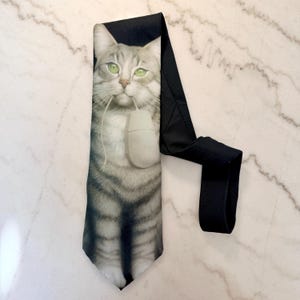 May include: A black necktie featuring a detailed image of a gray tabby cat holding a computer mouse in its mouth. The cat has green eyes and is set against a black background. The tie is made of a smooth fabric.