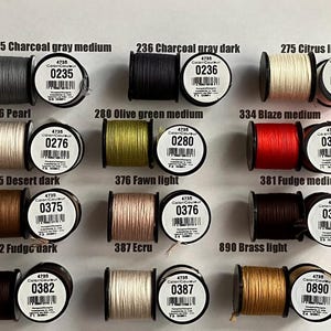 May include: A collection of spools of thread in various colors, including gray, black, olive green, red, brown, and gold. Each spool has a label with a color number and name, such as "235 Charcoal gray medium" and "275 Citrus light".
