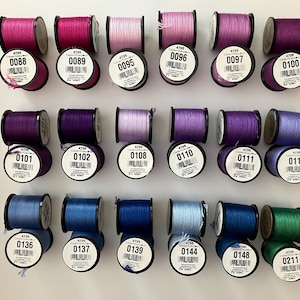 May include: Eighteen spools of embroidery thread arranged in three rows of six. The spools feature various shades of pink, purple, blue, and green. Each spool has a white circular label with a black barcode and a unique color code number.