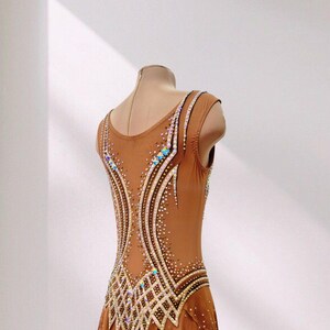 May include: A tan sleeveless leotard featuring a scoop neckline and a sheer skirt overlay. The bodice is decorated with intricate patterns of clear and iridescent crystals arranged in curved lines and geometric shapes across the back.
