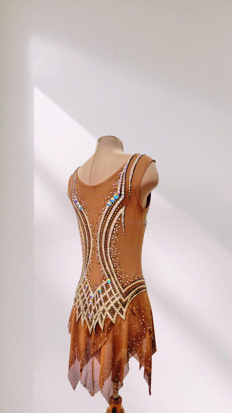 May include: A tan sleeveless leotard featuring a scoop neckline and a sheer skirt overlay. The bodice is decorated with intricate patterns of clear and iridescent crystals arranged in curved lines and geometric shapes across the back.