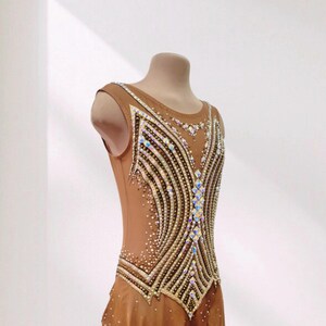 May include: A sleeveless tan leotard featuring a complex pattern of gold and black lines across the bodice, embellished with numerous iridescent crystals. The garment includes a sheer, layered skirt section adorned with scattered small crystals.