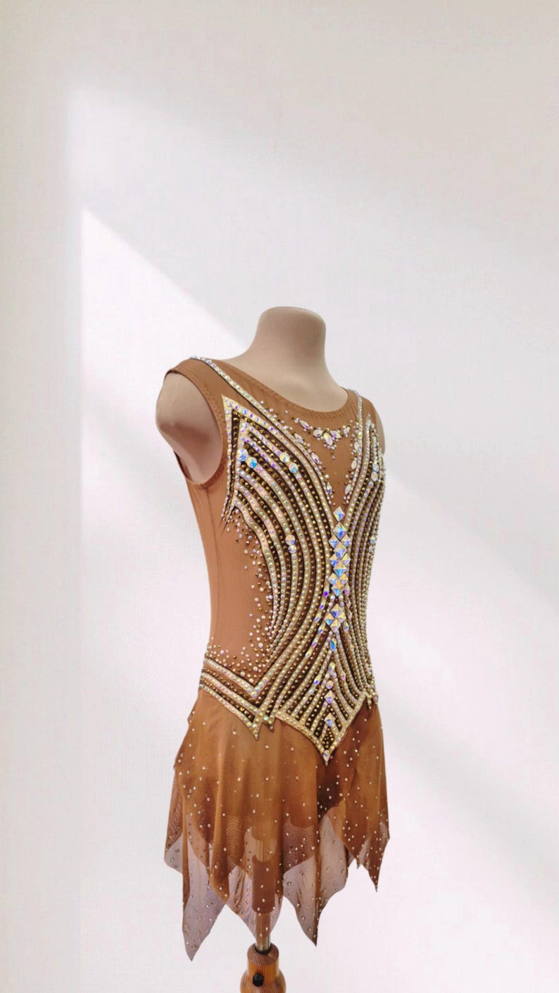 May include: A sleeveless tan leotard featuring a complex pattern of gold and black lines across the bodice, embellished with numerous iridescent crystals. The garment includes a sheer, layered skirt section adorned with scattered small crystals.