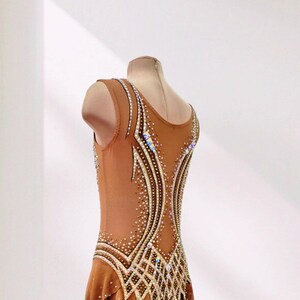 May include: A sleeveless tan leotard featuring a scoop neckline and a flowing skirt. The garment is decorated with intricate patterns of clear and iridescent crystals arranged in curved lines along the bodice and skirt, adding texture and shimmer.