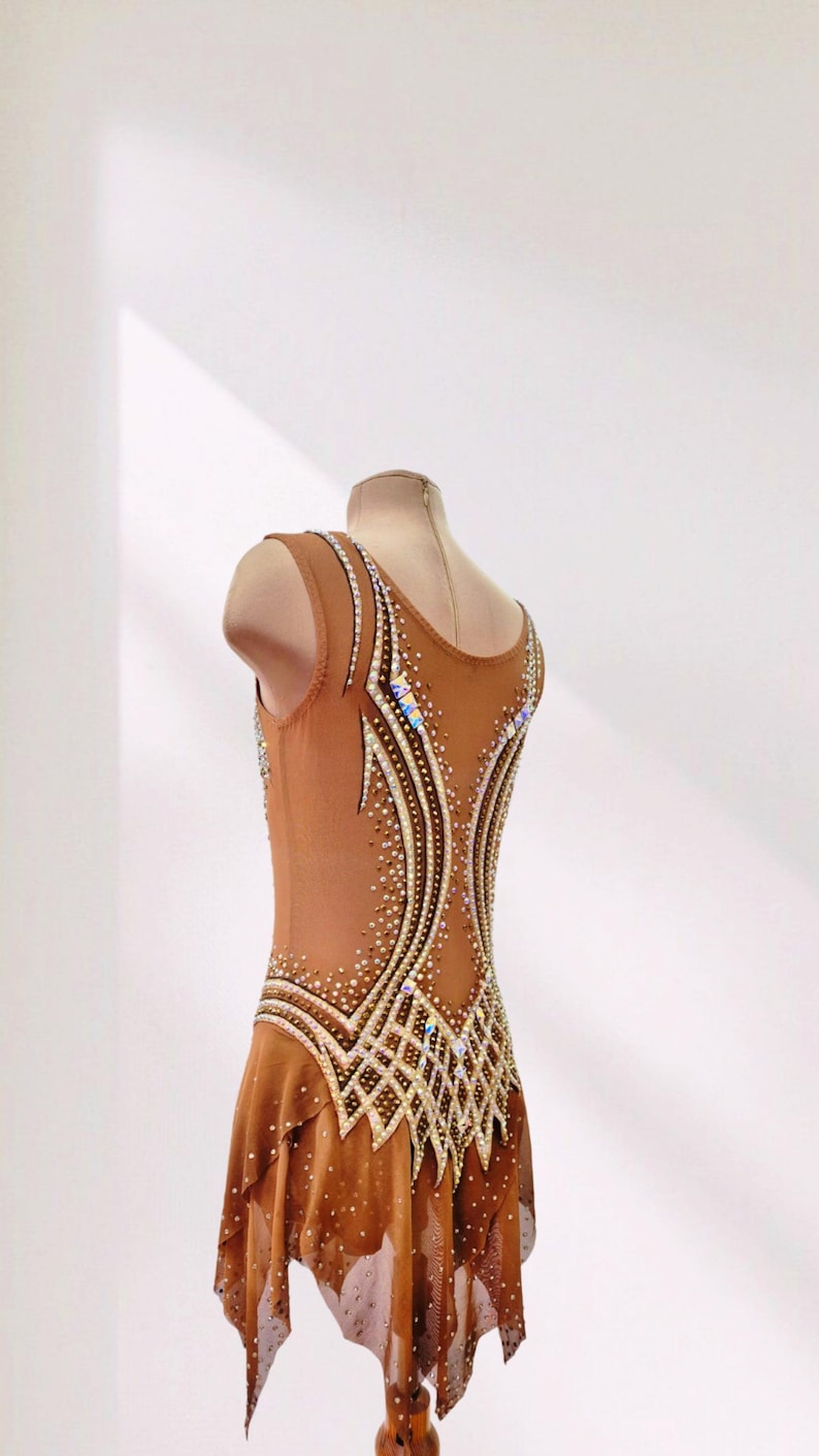 May include: A sleeveless tan leotard featuring a scoop neckline and a flowing skirt. The garment is decorated with intricate patterns of clear and iridescent crystals arranged in curved lines along the bodice and skirt, adding texture and shimmer.