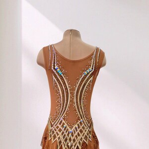 May include: A sleeveless tan leotard featuring a scoop neckline and a layered skirt. The bodice is decorated with intricate patterns of gold beads and iridescent crystals arranged in curved lines and a lattice design across the lower torso.