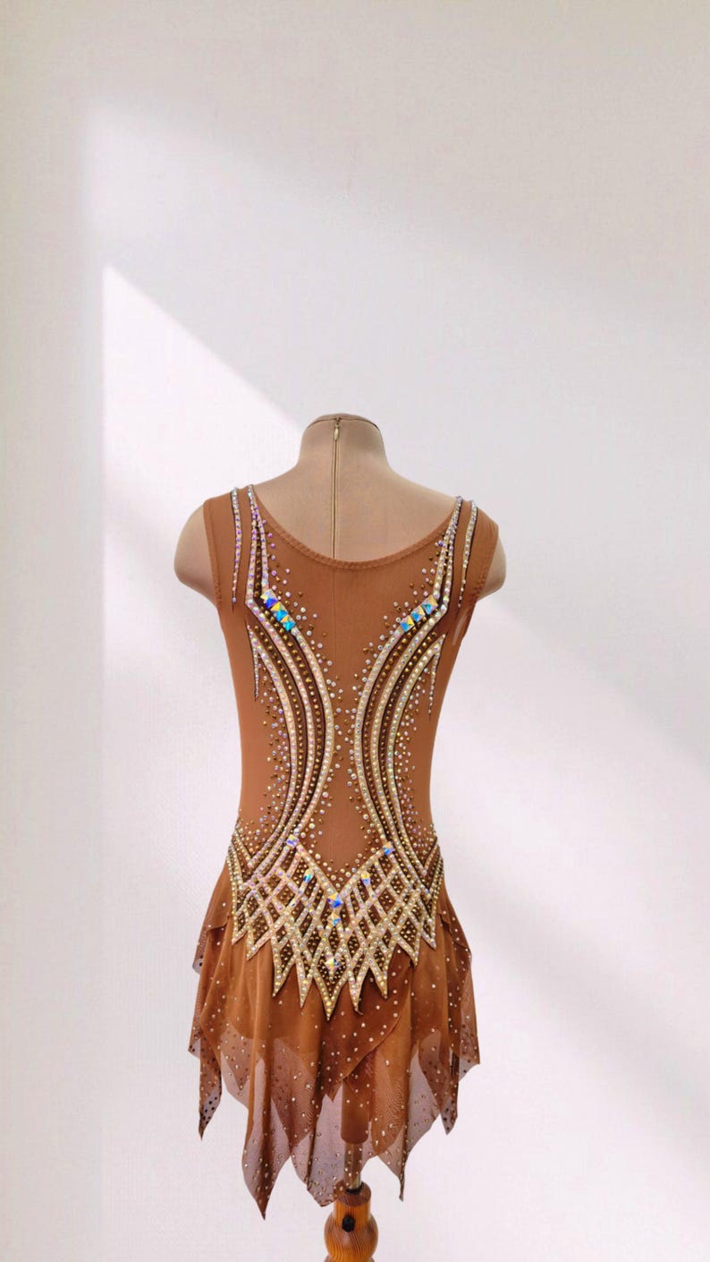 May include: A sleeveless tan leotard featuring a scoop neckline and a layered skirt. The bodice is decorated with intricate patterns of gold beads and iridescent crystals arranged in curved lines and a lattice design across the lower torso.
