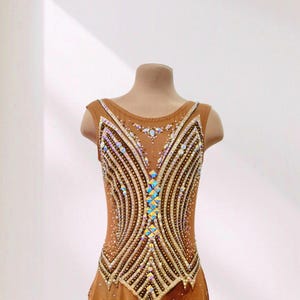 May include: A tan sleeveless leotard featuring a complex pattern of iridescent crystals and beads arranged in vertical lines. The bodice has a central column of diamond-shaped stones, while the skirt is made of sheer fabric with scattered crystals.