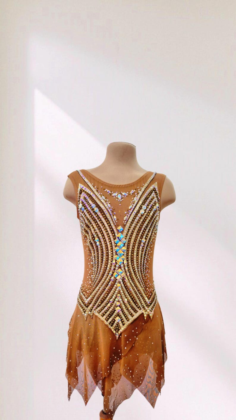 May include: A tan sleeveless leotard featuring a complex pattern of iridescent crystals and beads arranged in vertical lines. The bodice has a central column of diamond-shaped stones, while the skirt is made of sheer fabric with scattered crystals.