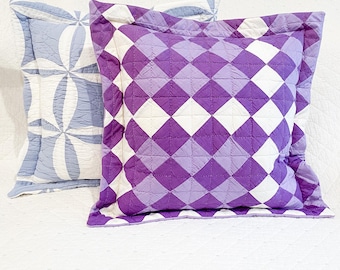 May include: Two square quilted pillows. The front pillow features a purple and white diamond grid pattern. The pillow behind it displays a light blue and white curved geometric design. Both pillows have a quilted texture and a flanged edge.