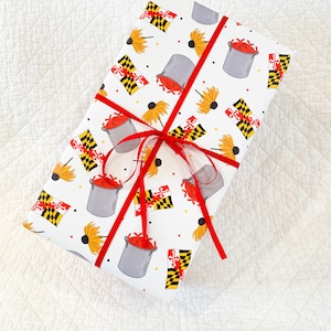 May include: A wrapped gift with white paper featuring a pattern of yellow flowers, Maryland flags, and gray pots filled with red crabs. The gift is tied with a bright red ribbon and bow, set against a white quilted background.