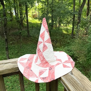 May include: A conical hat constructed from quilted fabric featuring a patchwork design of white and coral pink sections. The fabric displays a repeating floral pattern in dark gray and white, with a wide, circular brim and a pointed, upright crown.