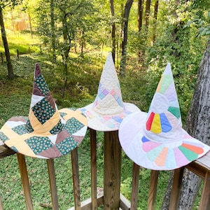 May include: Three quilted cone-shaped hats with wide brims. The left hat features a dark green floral, brown, and yellow patchwork pattern. The center hat is cream with pastel patchwork squares. The right hat has a white base with a colorful petal design.