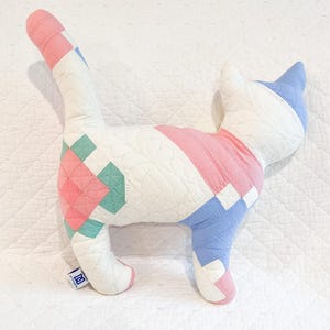 May include: A quilted cat-shaped pillow in a patchwork design. The pillow features a white base with pink, blue, and teal geometric patterns. The cat has a long tail and pointed ears. The pillow is made of soft fabric and has a stitched design.