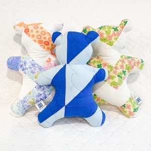 May include: Three stuffed bear-shaped toys made from quilted fabric patches. The center bear is blue and light blue. The left bear features orange honeycomb and purple floral patterns. The right bear has a green and pink clover print pattern.