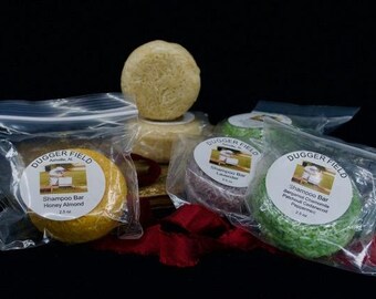 May include: A collection of shampoo bars in clear plastic bags, each with a label. The bars are various colors, including yellow, green, and purple. The labels read "Dugger Field" and list the scent and weight, 2.5 oz.