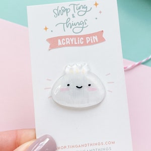 May include: White acrylic pin featuring a cartoon dumpling design. The dumpling has a smiling face and a pink blush. The pin is displayed on a white card with the text "Shop Ting & Things Acrylic Pin" and the website address "shop.tingandthings.com".