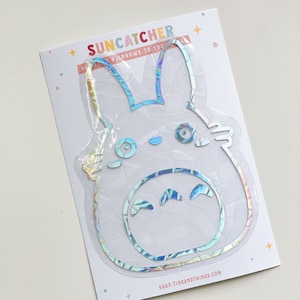 May include: A holographic sticker of a cartoon character with large ears and a smiling face. The sticker is on a white card with the text "Suncatcher" and "Send rainbows to your...