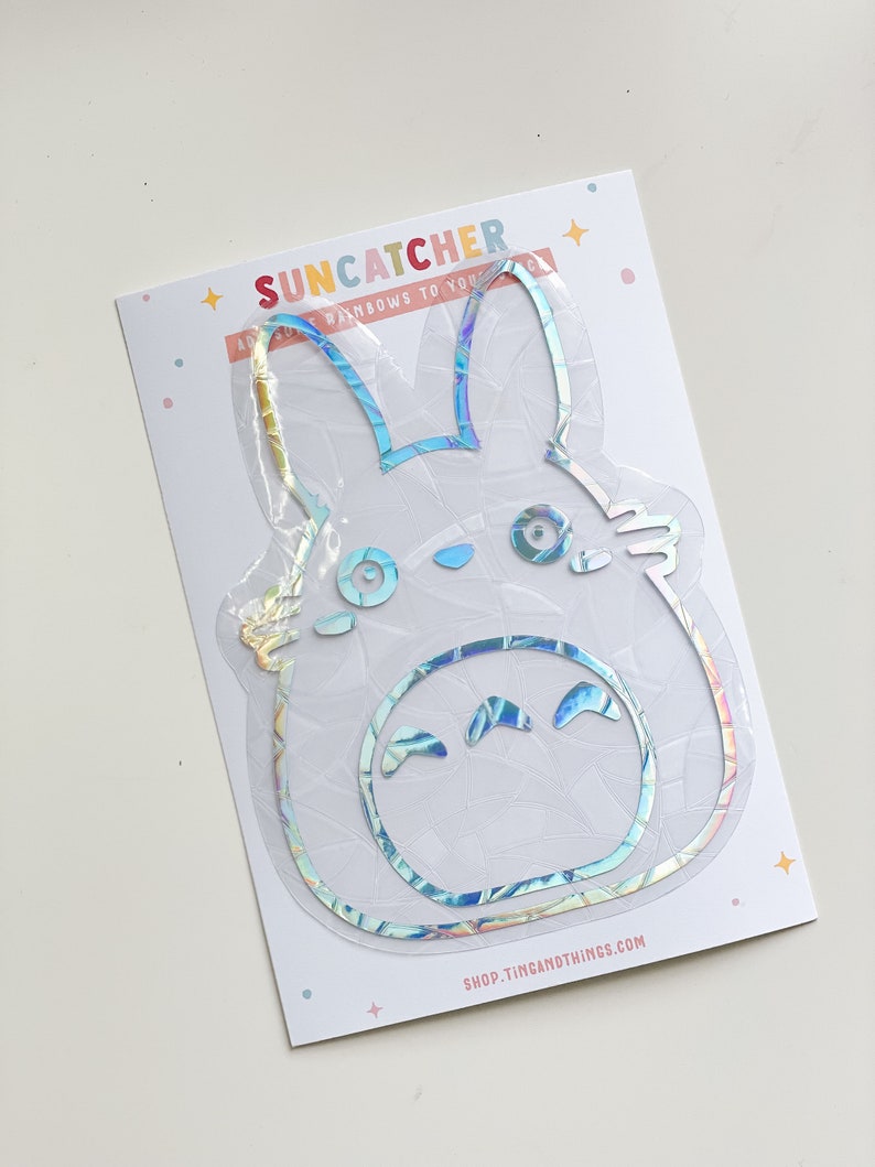 May include: A holographic sticker of a cartoon character with large ears and a smiling face. The sticker is on a white card with the text "Suncatcher" and "Send rainbows to your...