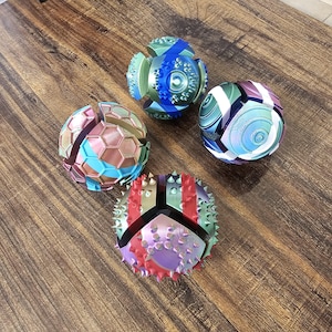 May include: Four colorful, geometric puzzle balls rest on a wooden surface. The spheres feature various designs, including spikes, hexagons, and striped patterns. Colors include blue, green, red, and purple. The balls appear to be 3D-printed.